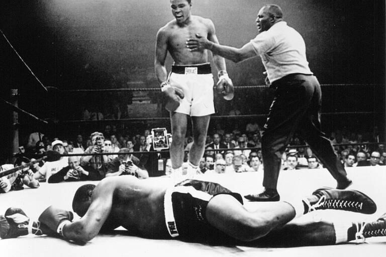 Mohamed Ali sur le ring en noir et blanc avec un adversaire K.O à ses pieds et l'arbitre qui vient l'arrêter. 