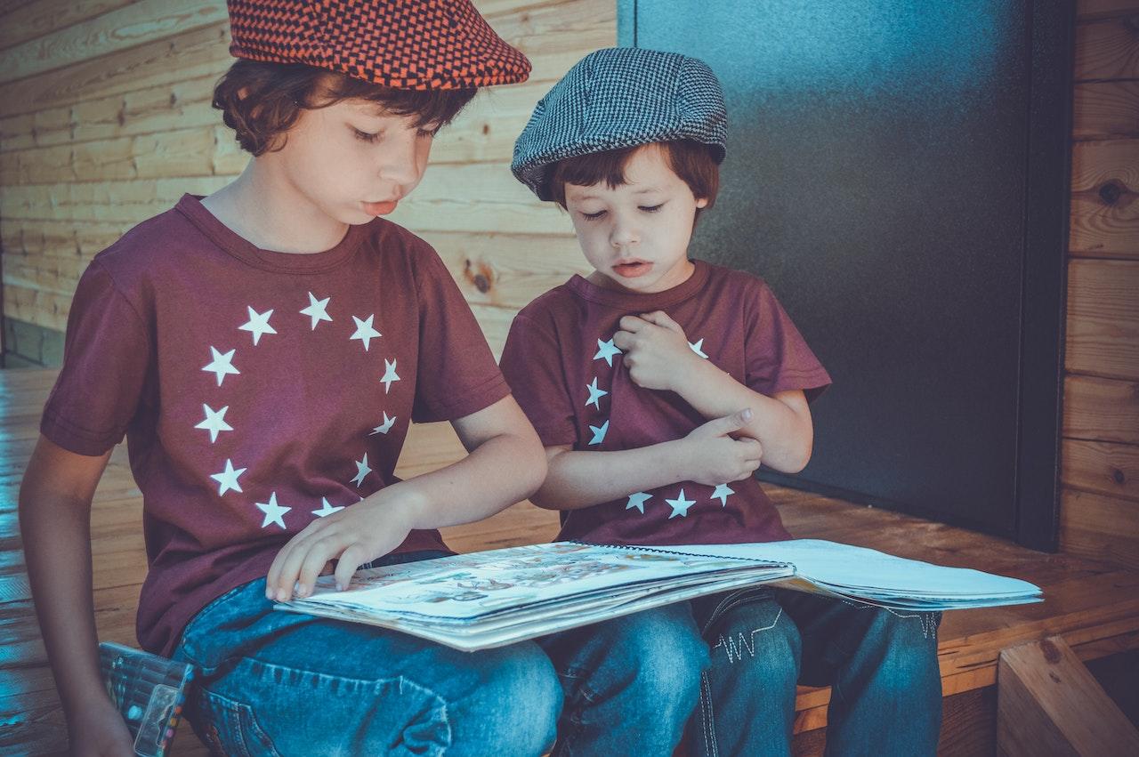 Un grand frère de 7-8 ans lit une histoire à son petit frère de 4-5 ans. Ils sont habillés pareil et assis sur une marche en bois. 