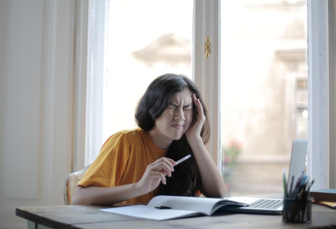 Jeune fille d'environ 14-15 ans qui se tient la tête. Elle est assise devant une table, un ordinateur ouvert, un cahier sous les yeux et un stylo dans la main. 