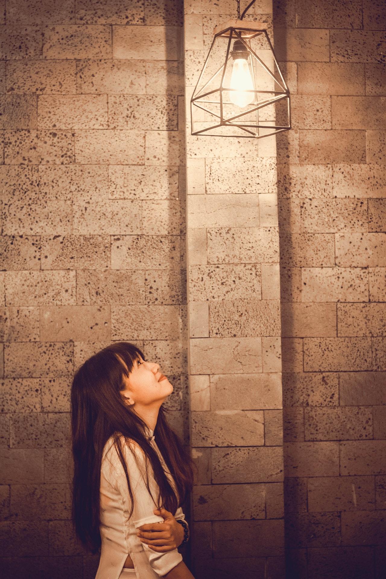 Jeune fille asiatique qui regarde une ampoule au plafond devant un mur en pierres grises
