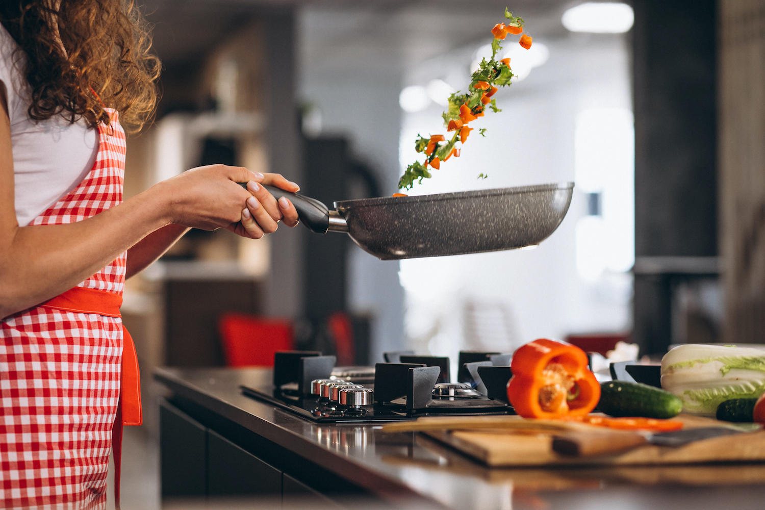 Femme avec tablier en train de faire revenir des légumes dans une cuisine.