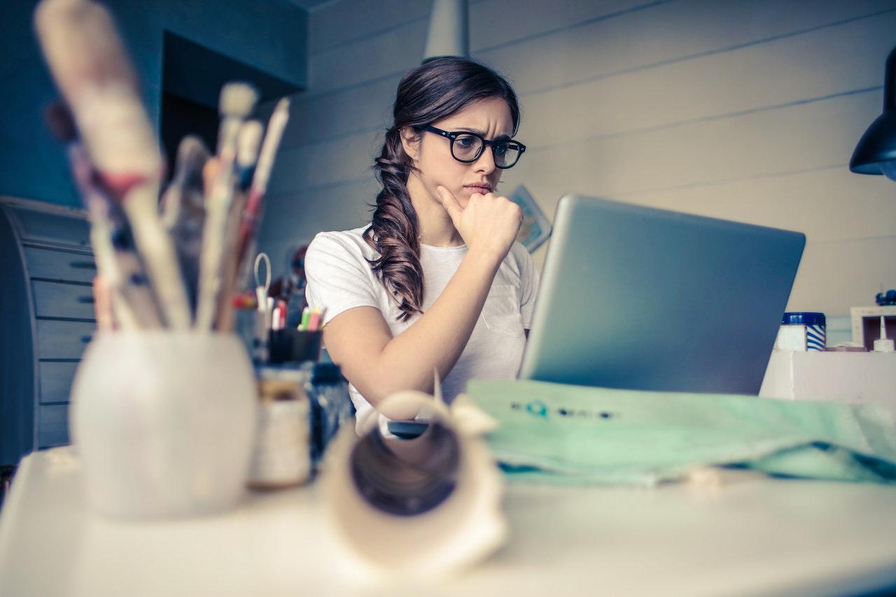 Jeune fille blanche assise derrière un bureau devant son ordinateur. Elle porte des lunettes et semble concentrée.