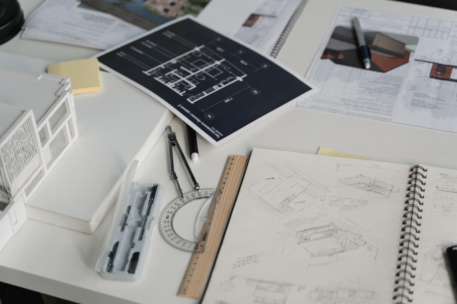Un carnet de croquis est ouvert près d'un plan, d'un compas, d'un rapporteur et d'une règle. 