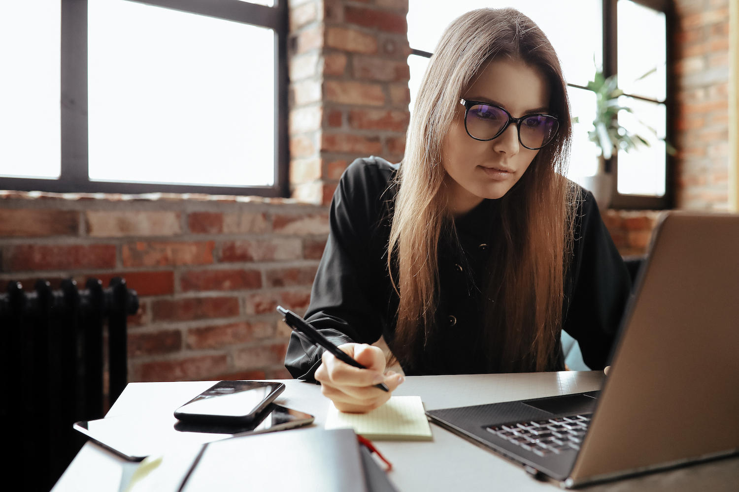 Jeune femme avec des lunettes devant un ordinateur en train de prendre des notes à la main.