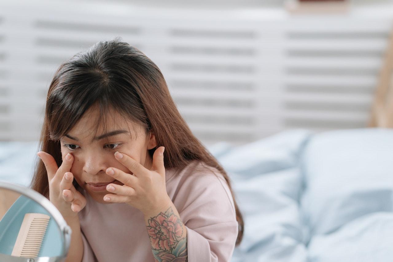 Jeune femme asiatique qui applique de la crème contour des yeux sur son visage en se regardant dans le miroir.
