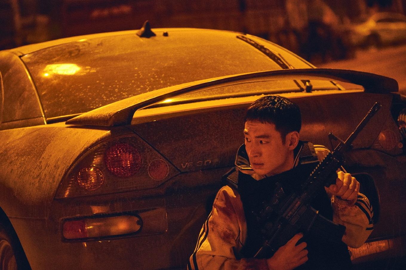 Un jeune homme est assis derrière une voiture avec une arme dans les mains.