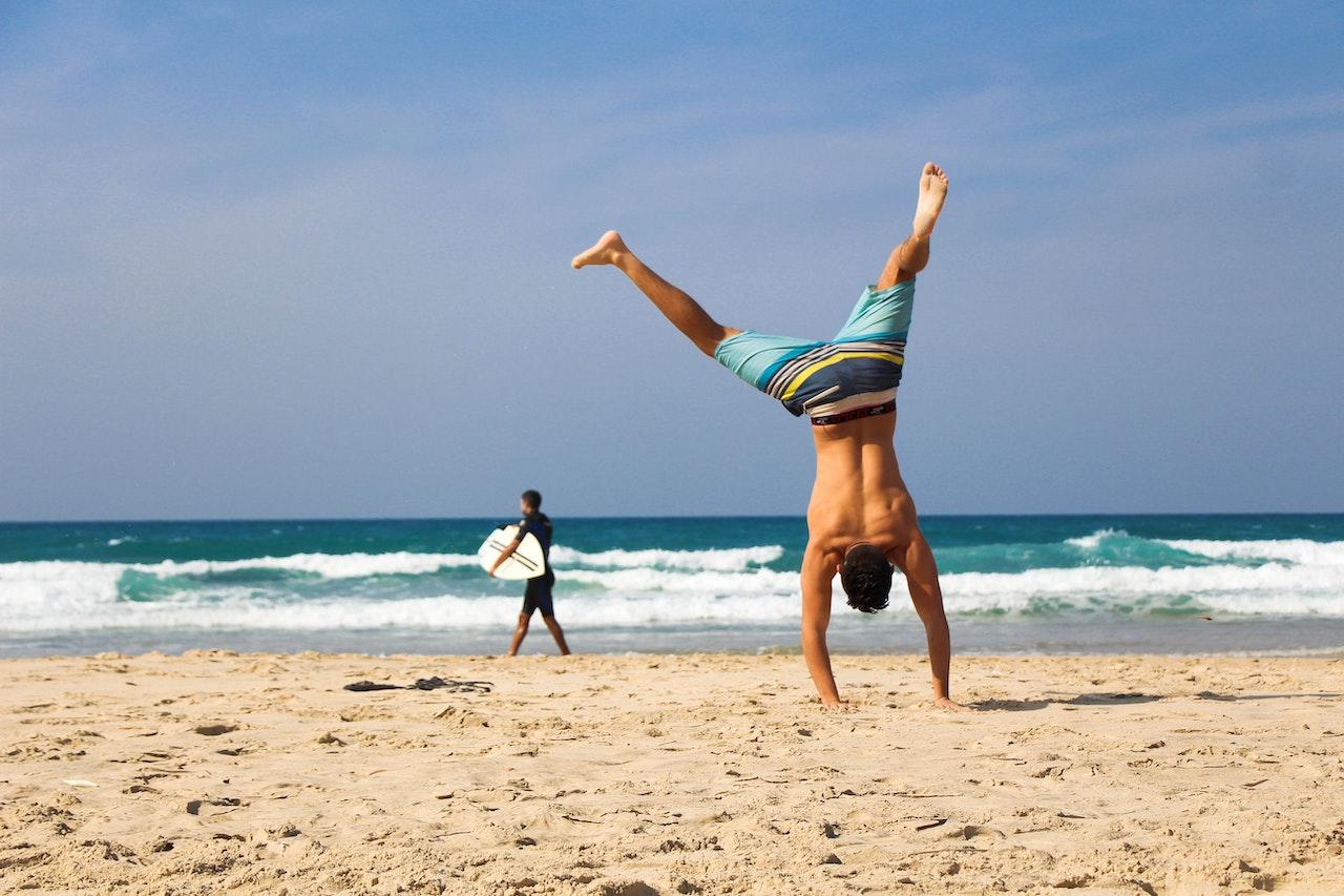 Homme de dos faisant une roue sur le sable, avec l'océan en fond et un autre homme qui marche en tenant une planche de surf. 