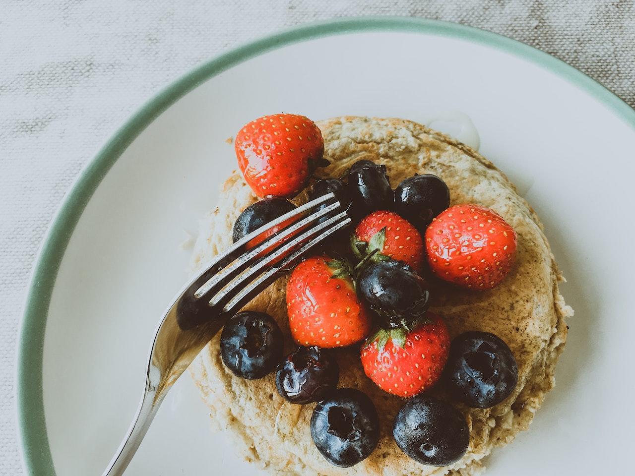 Un pancake posé dans une assiette avec des fraises et des myrtilles par dessus et une fourchette.