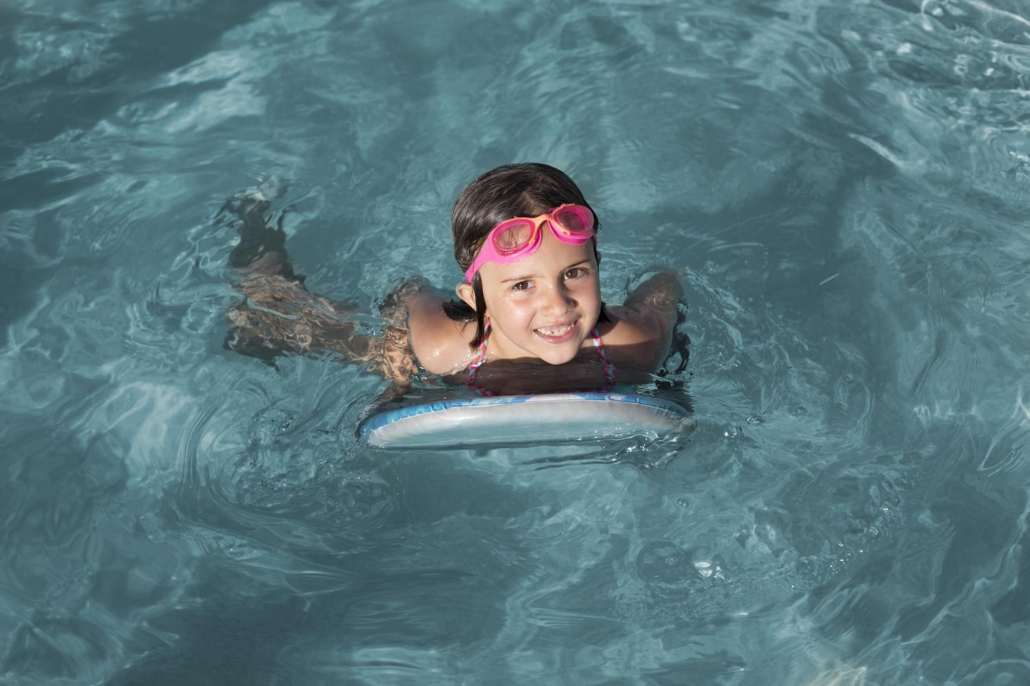 Une petite fille avec des lunettes de piscine sur la tête et une planche dans une piscine.