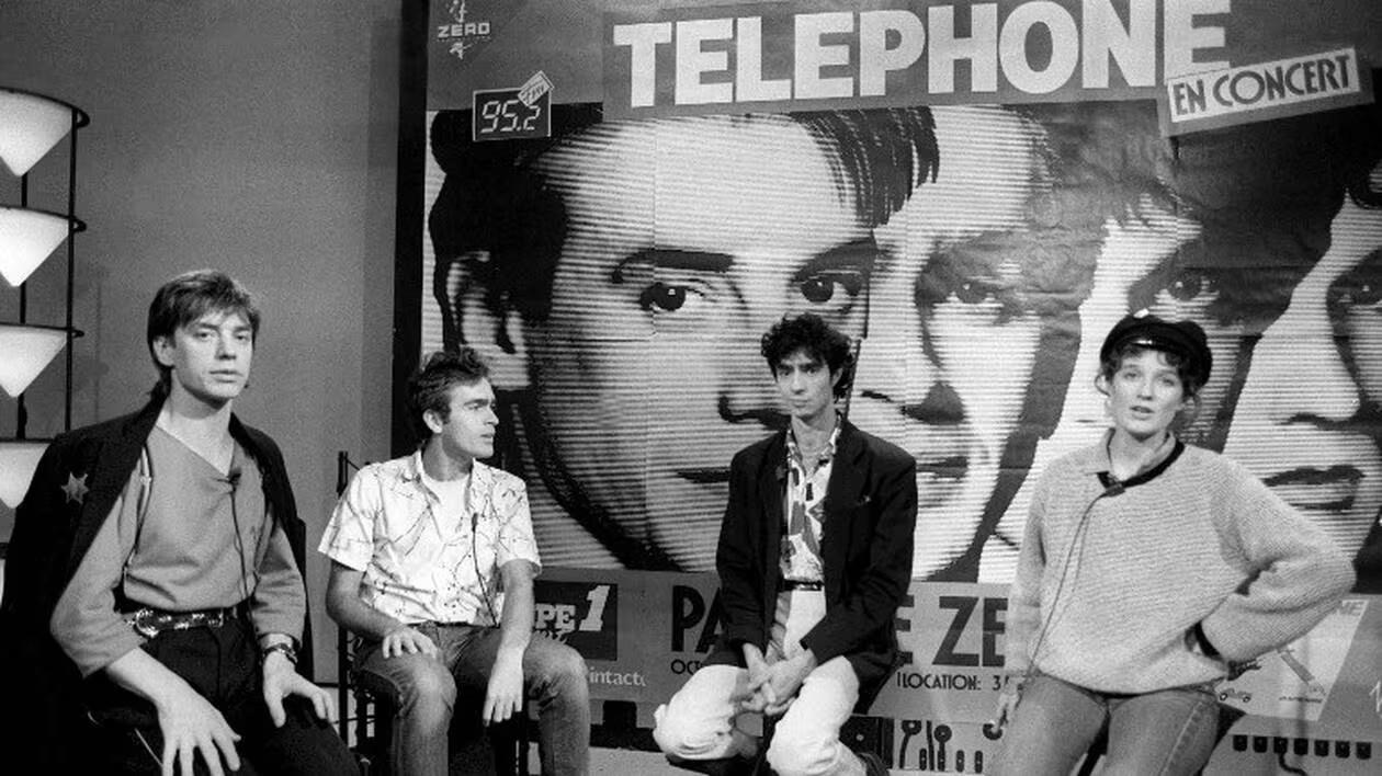 Les 4 membres du groupe Téléphone sont assis devant une affiche de concert.