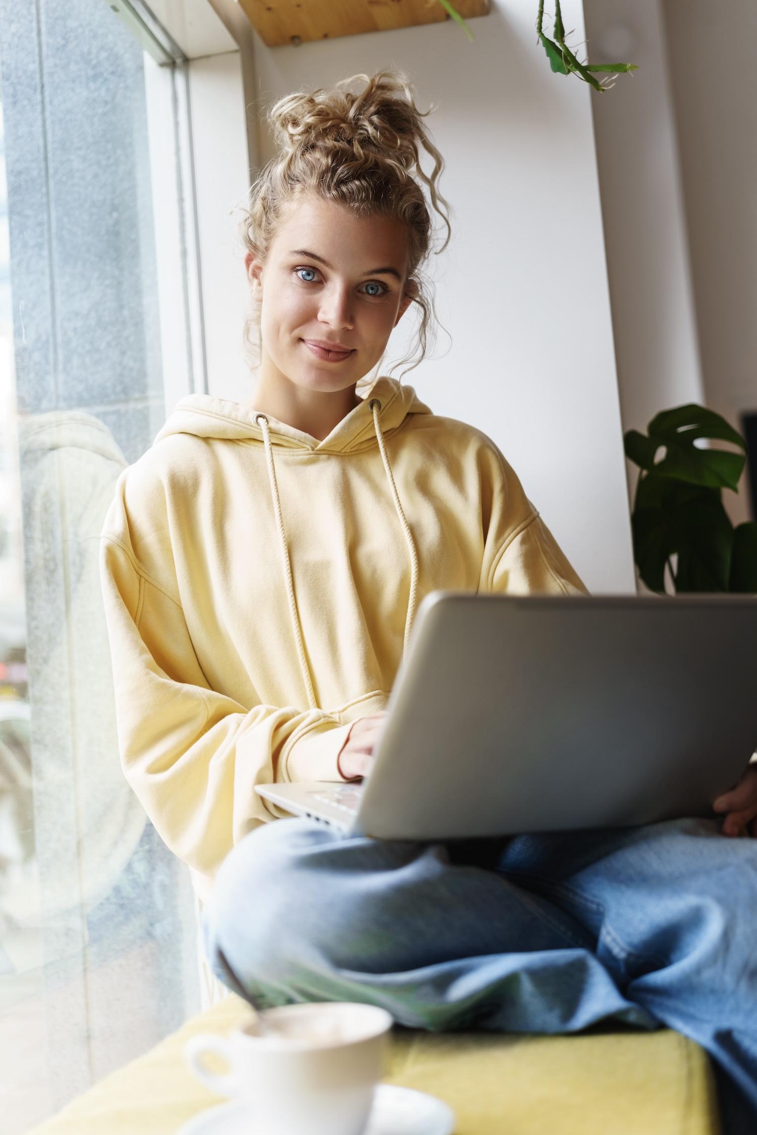 Jeune femme blonde assise sur le rebord d'une fenêtre avec un ordinateur sur les genoux.