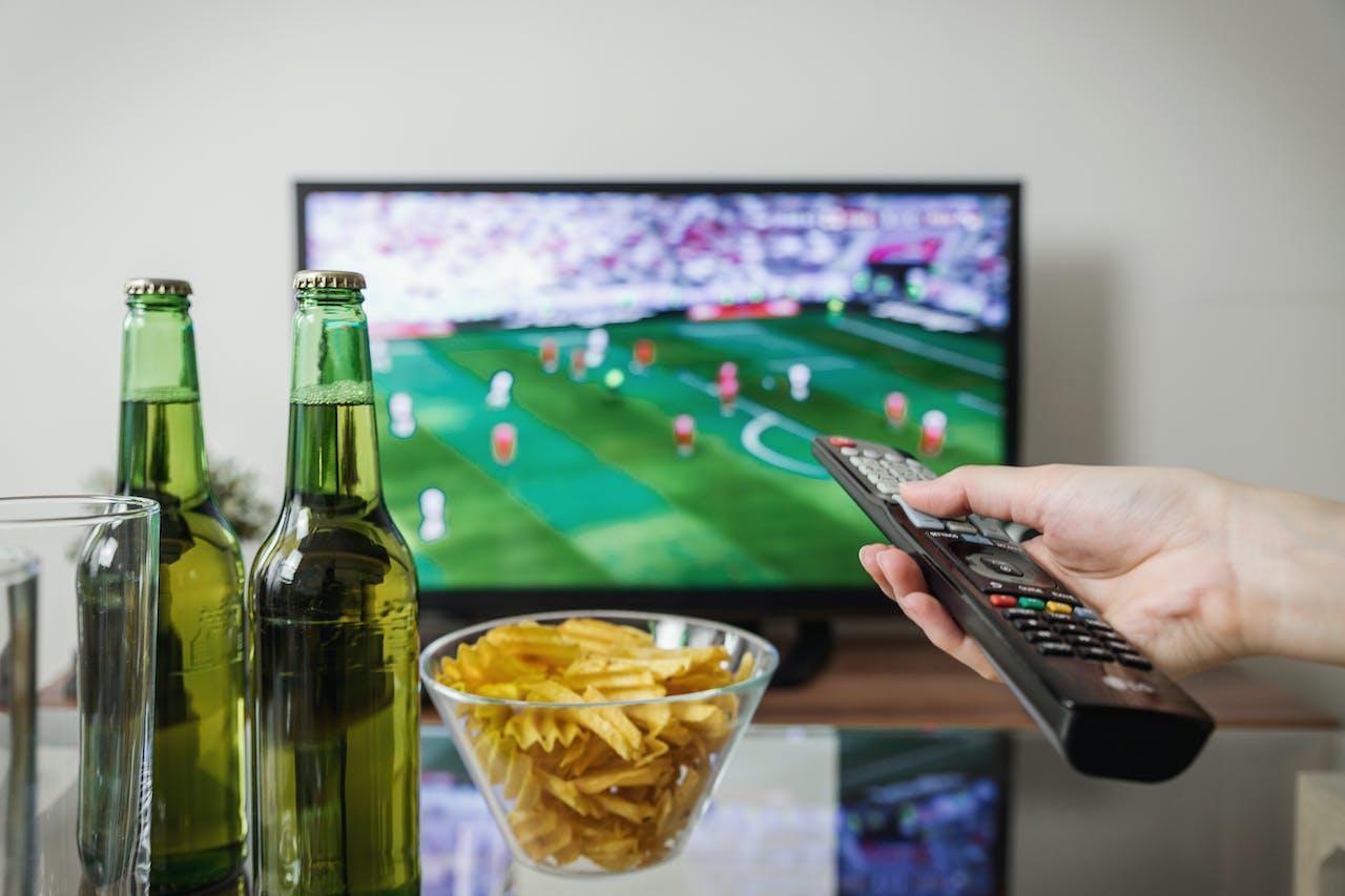 Match de foot à la télé avec quelqu'un qui a une télécommande à la main, deux bières sont posées sur la table basse avec de chips. 