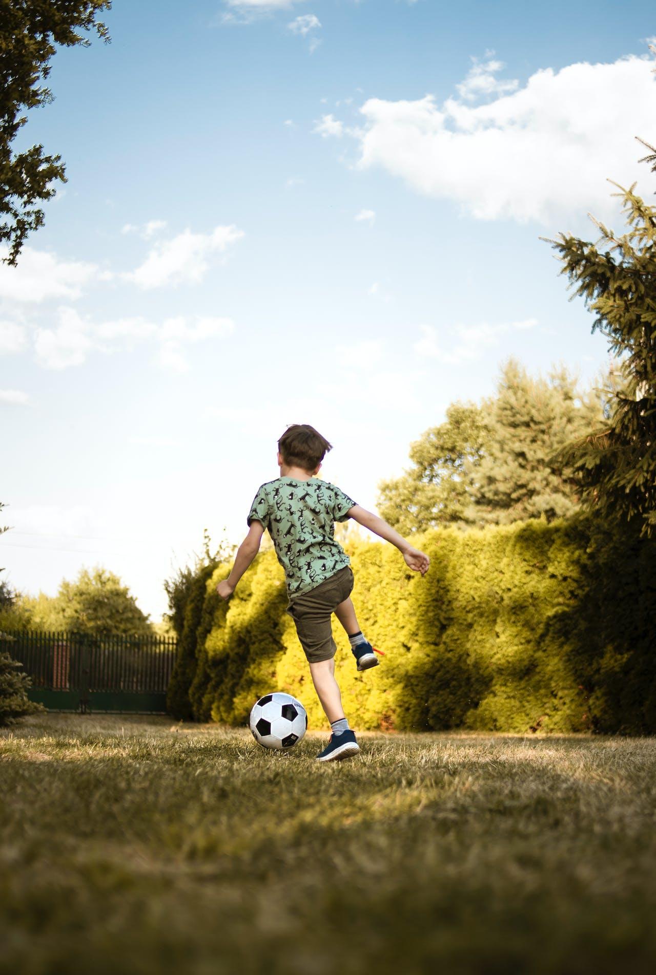 Jeune garçon de dos jouant avec un ballon de foot en extérieur.