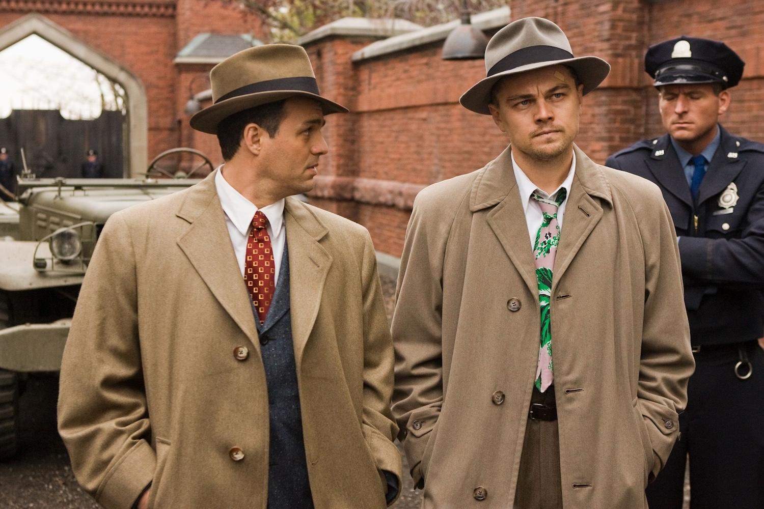 Deux hommes sont côte à côte en trench beige et avec un chapeau. 