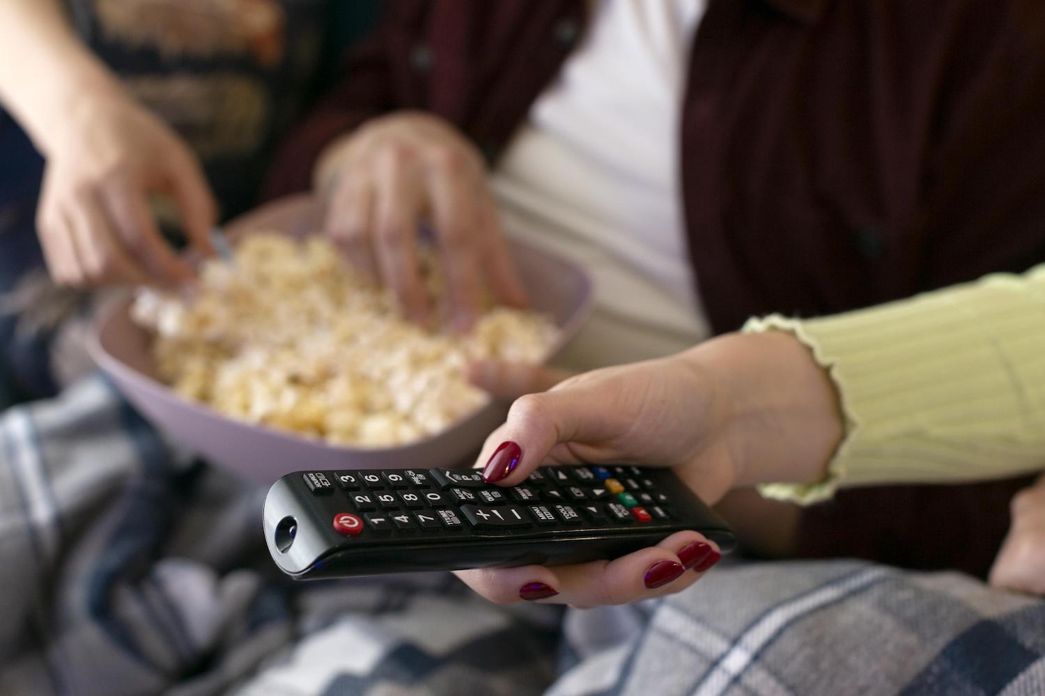 Une personne tient une télécommande et une autre un bol de popcorn.