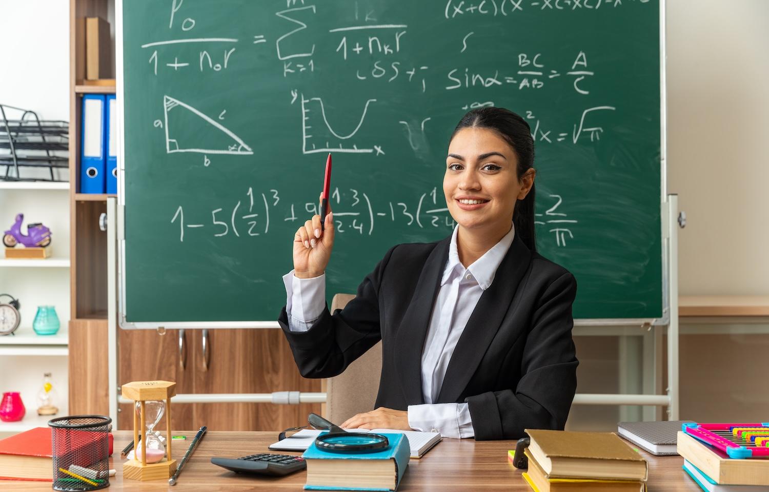 Une jeune professeur est assise à son bureau devant un tableau noir.