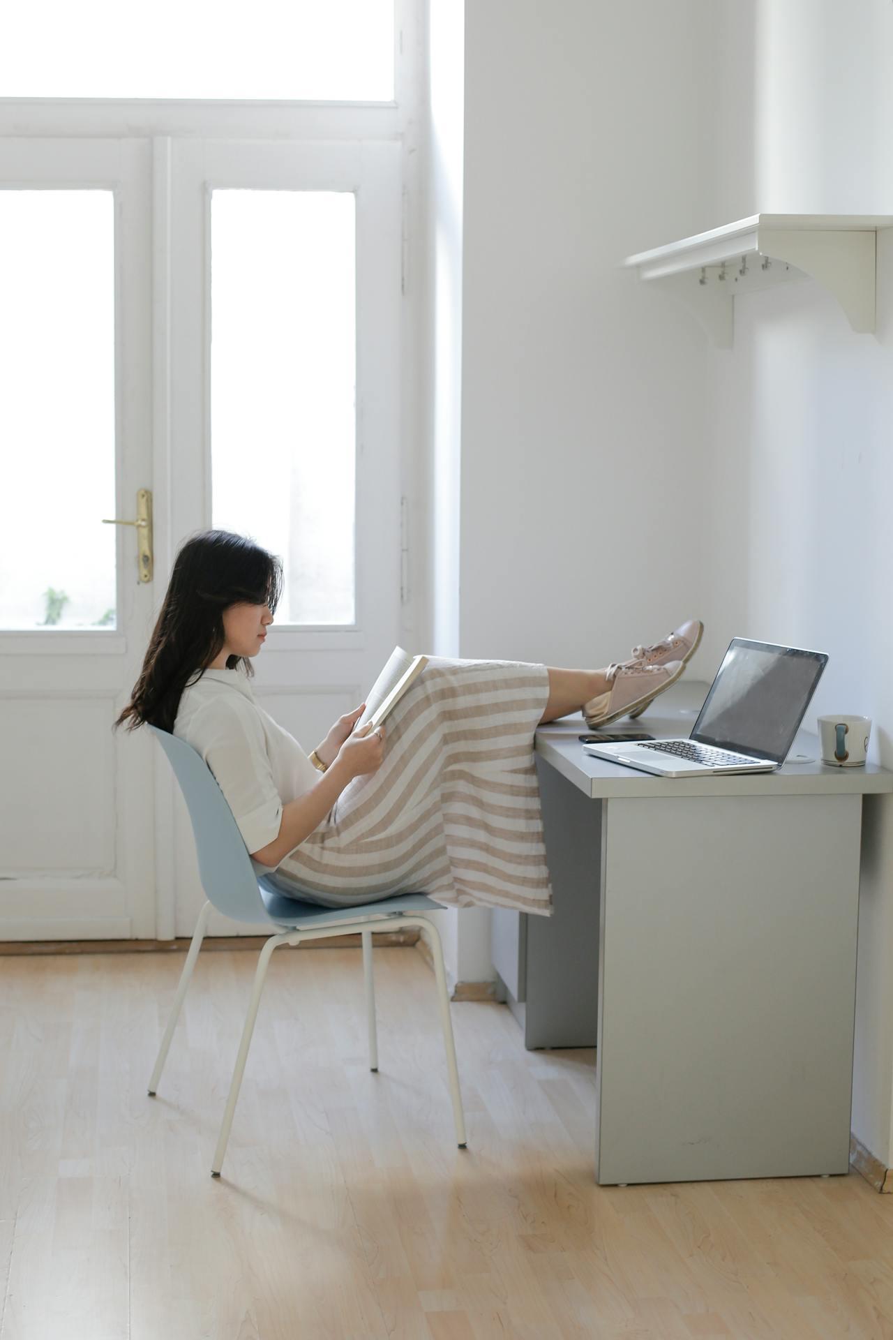 Jeune femme asiatique assise à son bureau, les pieds posés dessus en train de lire un livre devant son ordinateur. 