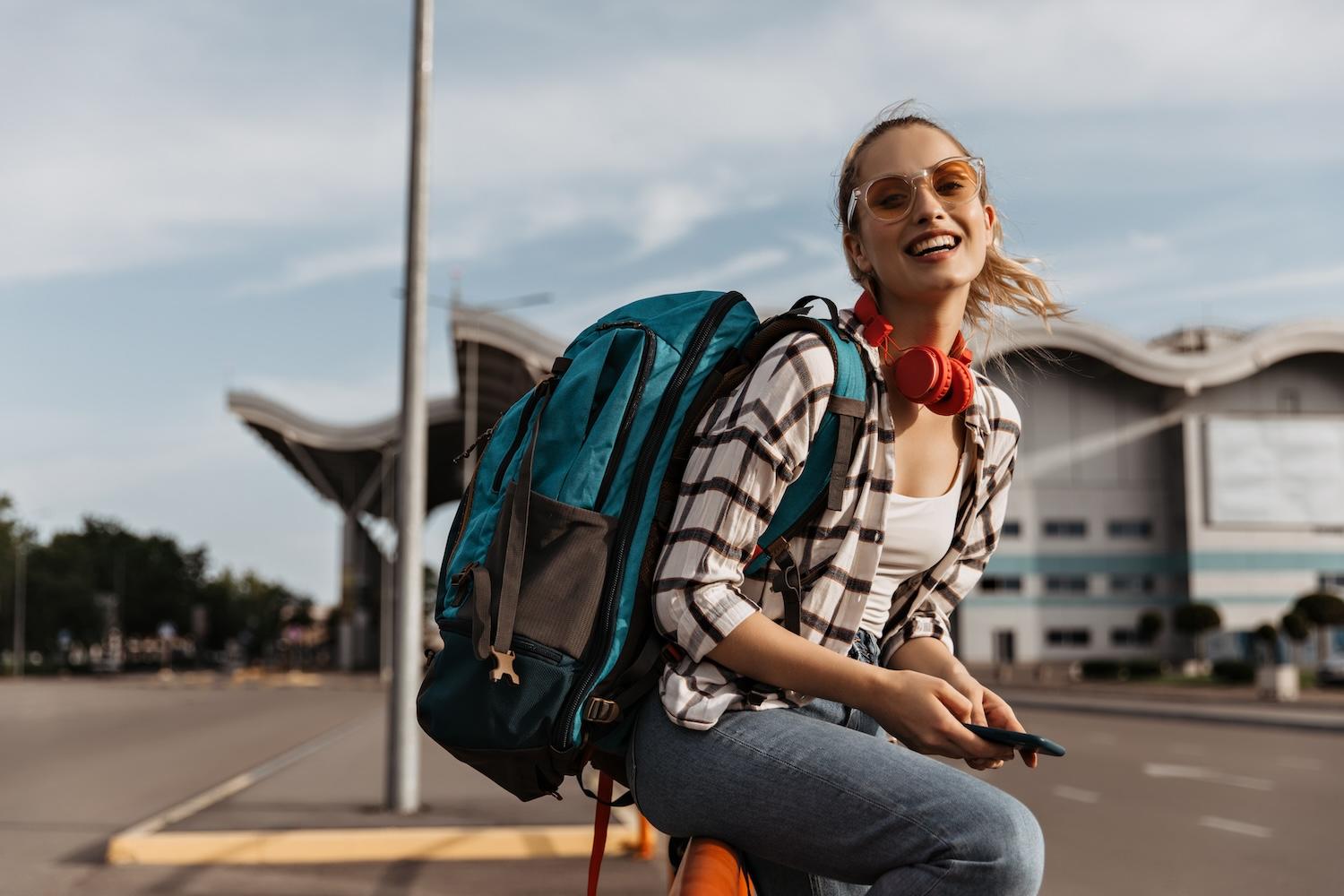 Une jeune femme est assise devant l'aéroport avec un sac sur le dos.