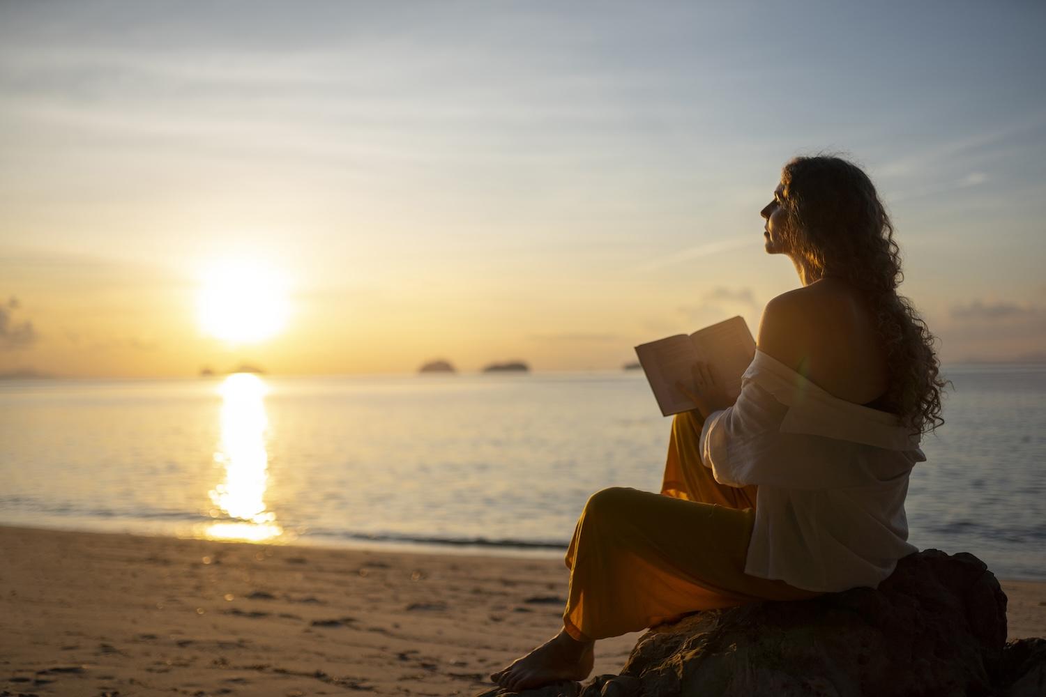 Une jeune femme est devant un coucher de soleil avec un livre.