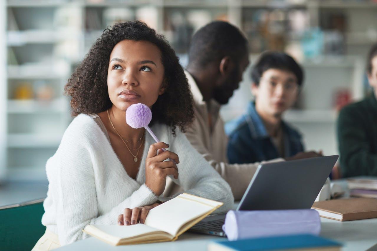 Jeune femme noire réfléchissant à son bureau, le stylo à la main, devant son ordinateur.