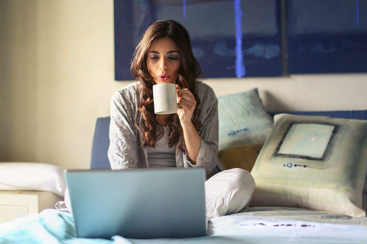 Jeune femme installée sur son lit devant son ordinateur avec une tasse de café dans la main.