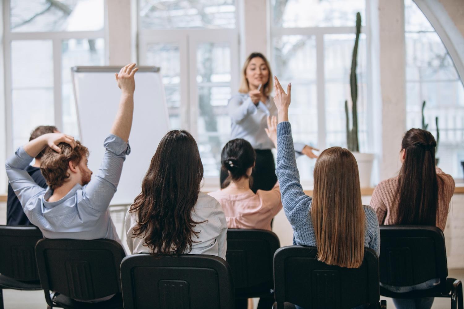 Plusieurs élèves de dos dans une classe qui lèvent le doigt.