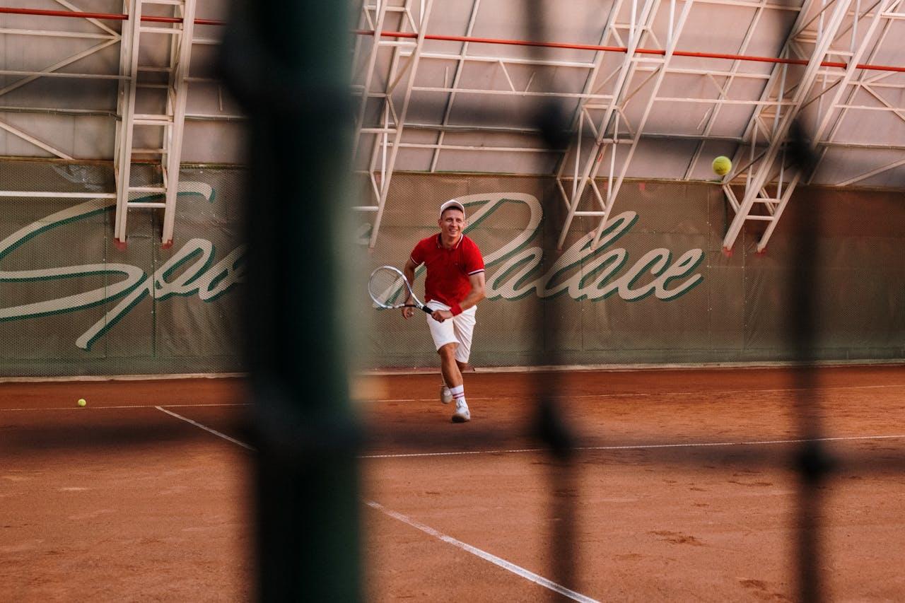 Homme courant sur un court de tennis en terre battue, on le voit à travers le filet.