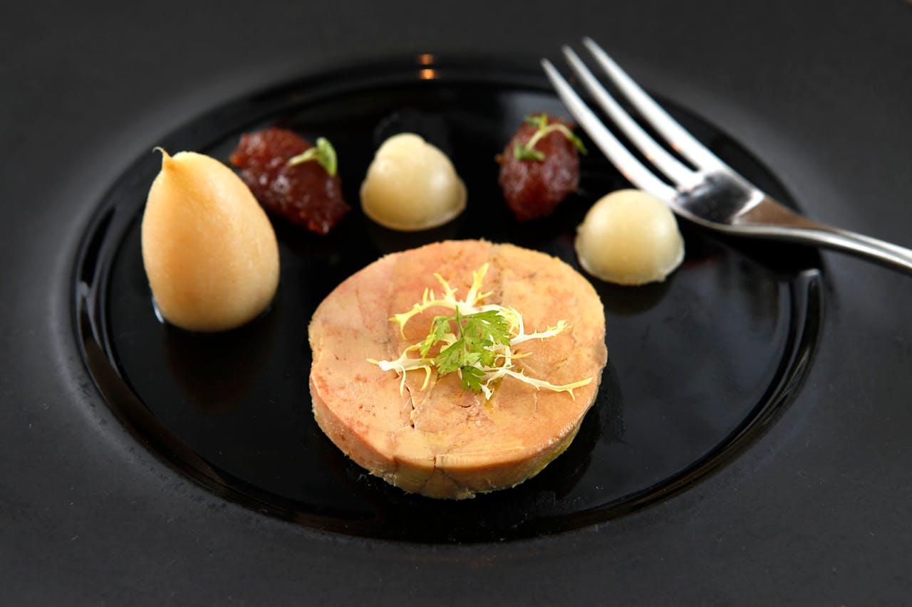 Tranche de foie gras posée sur une assiette noire avec une fourchette et des boules de confiture. 