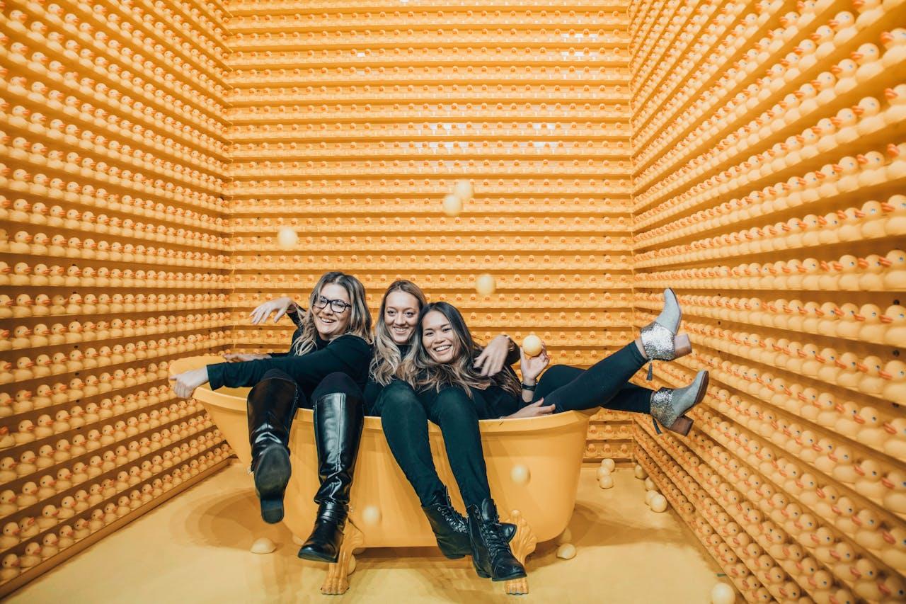 Trois jeunes femmes assises dans une baignoire au milieu de canards en plastique.