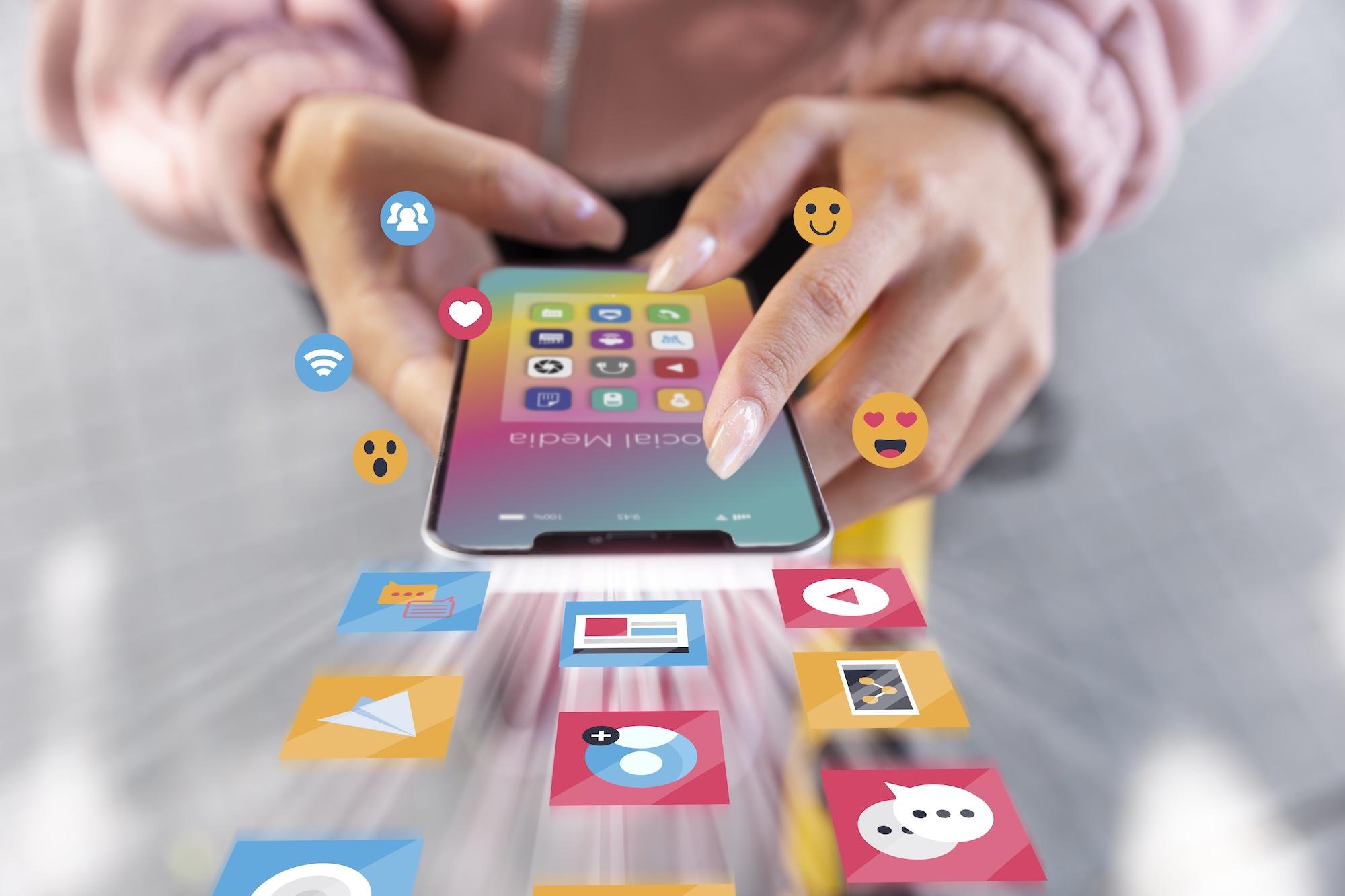 Une personne tient un smartphone dans ses mains avec des emojis et logos qui sortent du téléphone.