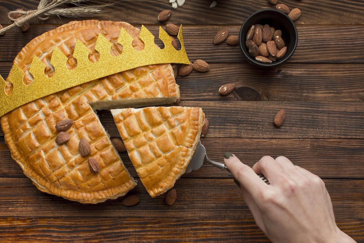 Une galette des rois vue d'en haut avec des amandes posées ça et là, une couronne dessus et une main qui vient prendre une part avec une pelle à tarte.