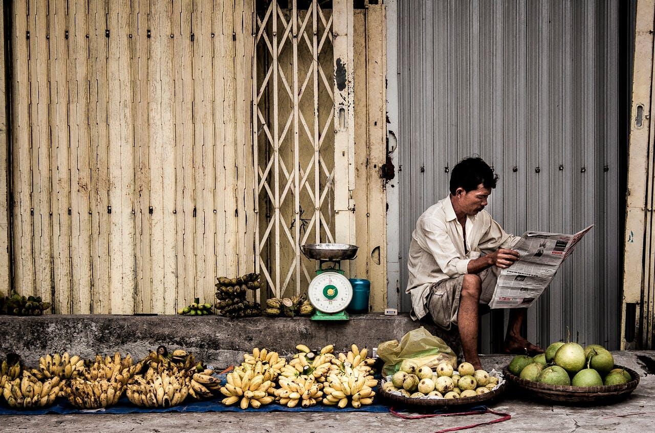 Homme vietnamien assis en train de lire son journal devant un étal de fruits. 