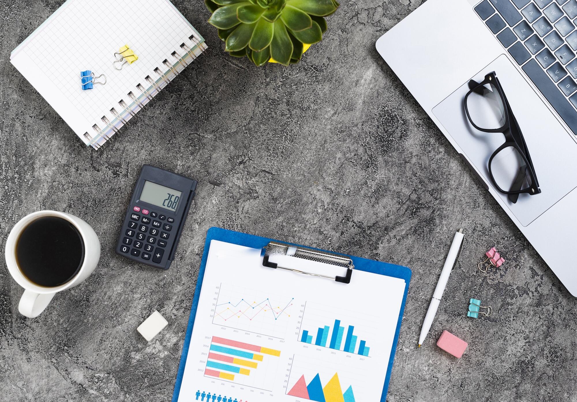 Un ordinateur est posé sur une table avec une calculette, un café et d'autres éléments de bureau.
