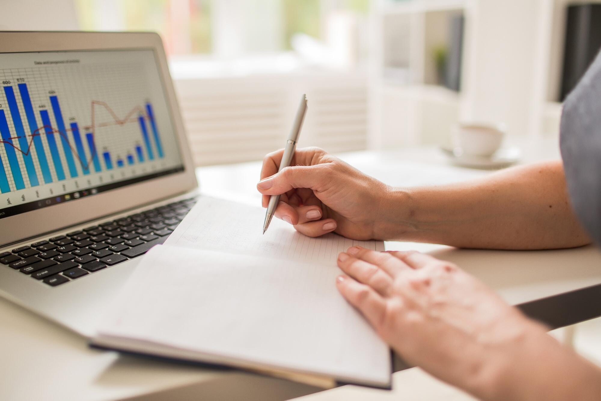 Une personne écrit sur un cahier avec des statistiques sur un ordinateur.
