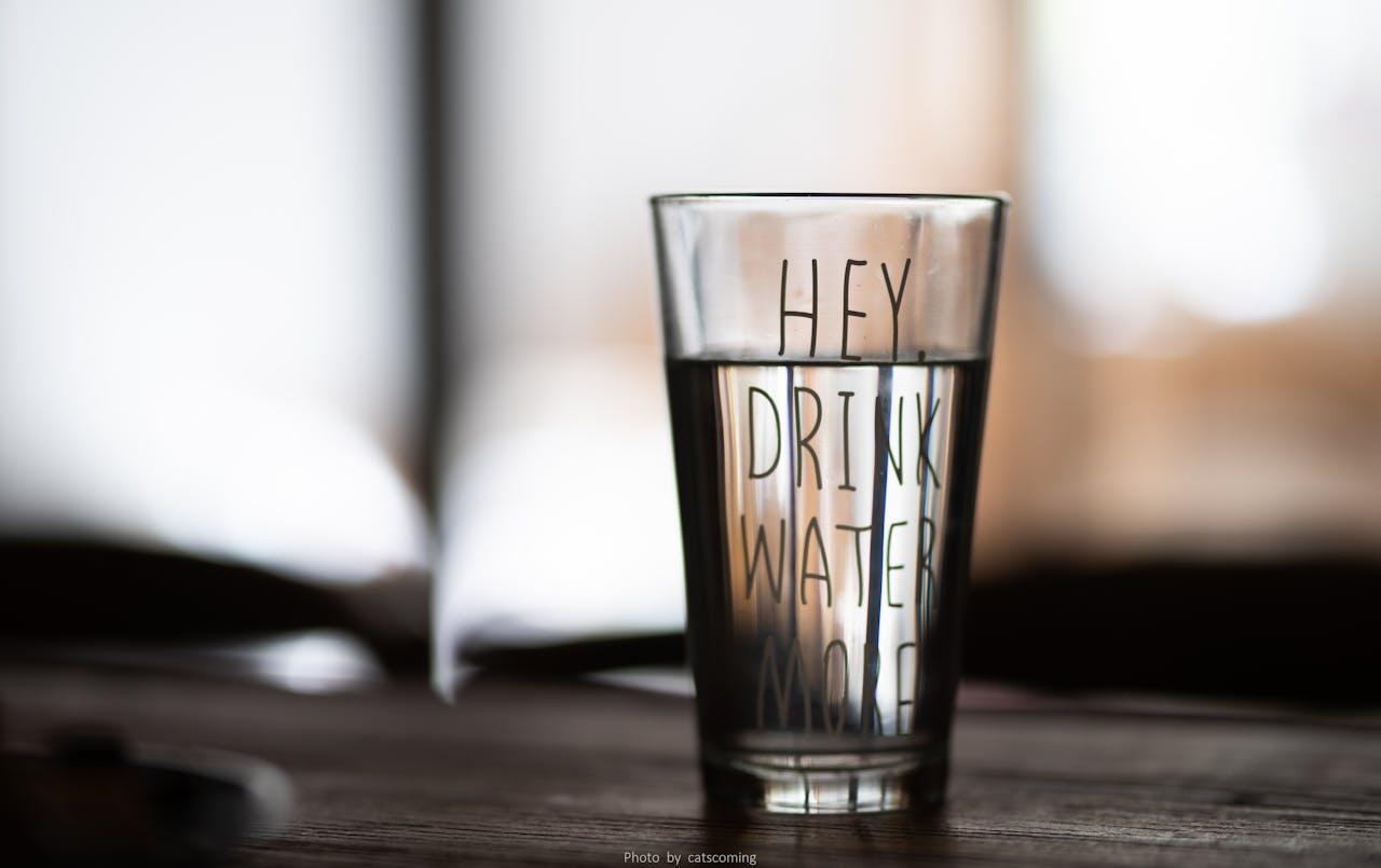 Verre d'eau posé sur une table avec écrit "Hey, drink water more" ce qui signifie "hé, bois plus d'eau". 