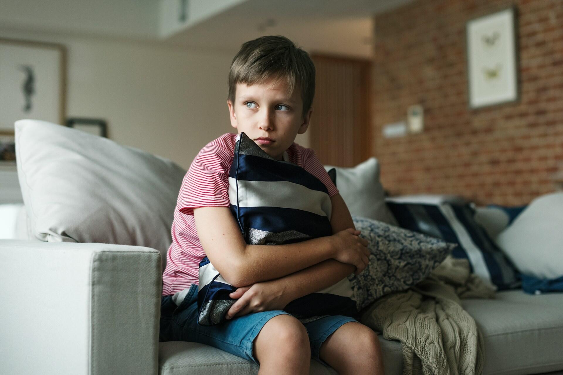 Un petit garçon qui a l'air triste sur un canapé et qui tient un coussin dans ses bras