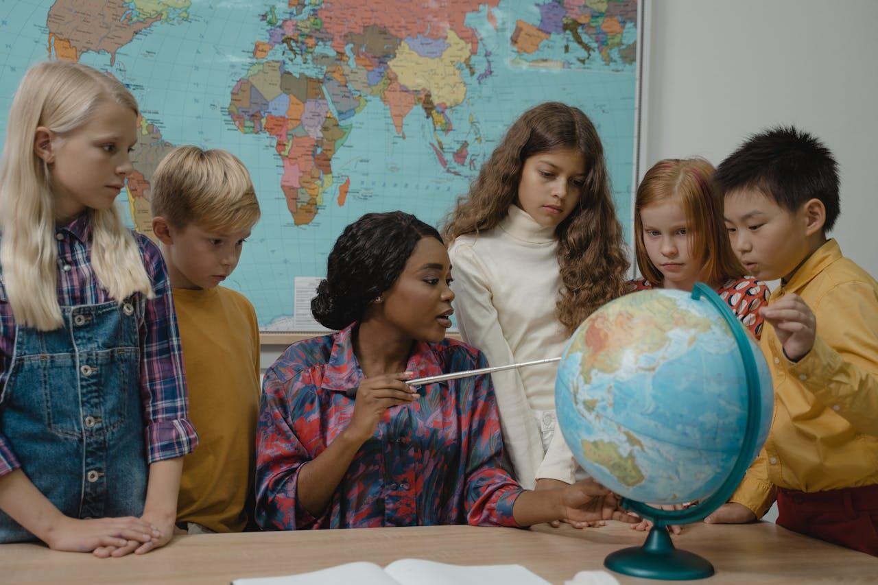 Jeune prof entourée d'élèves à son bureau et pointant un pays sur un globe terrestre. 