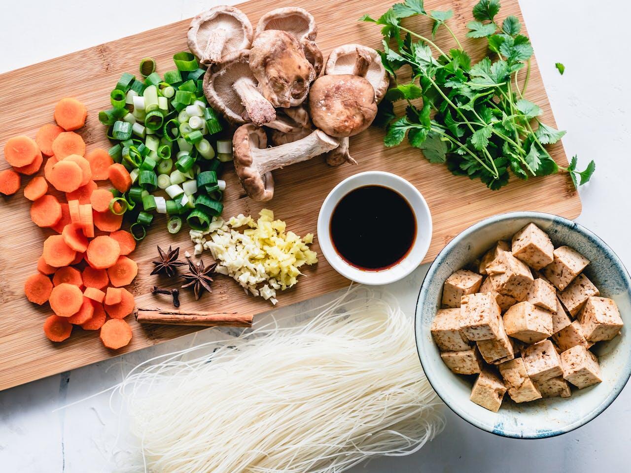 Planche à découper avec des légumes et un bol de tofu, ainsi que des nouilles de riz posées. 