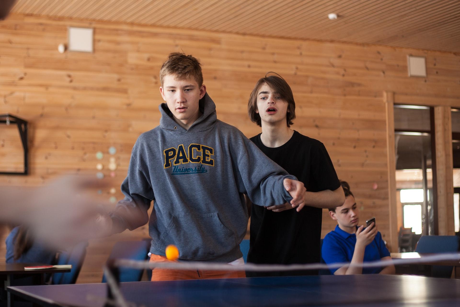 A boy in a grey hoodie plays ping pong while another boy in a black shirt stands behind him, looking on and a third boy in a blue shirt sits against the wall, looking at his mobile.