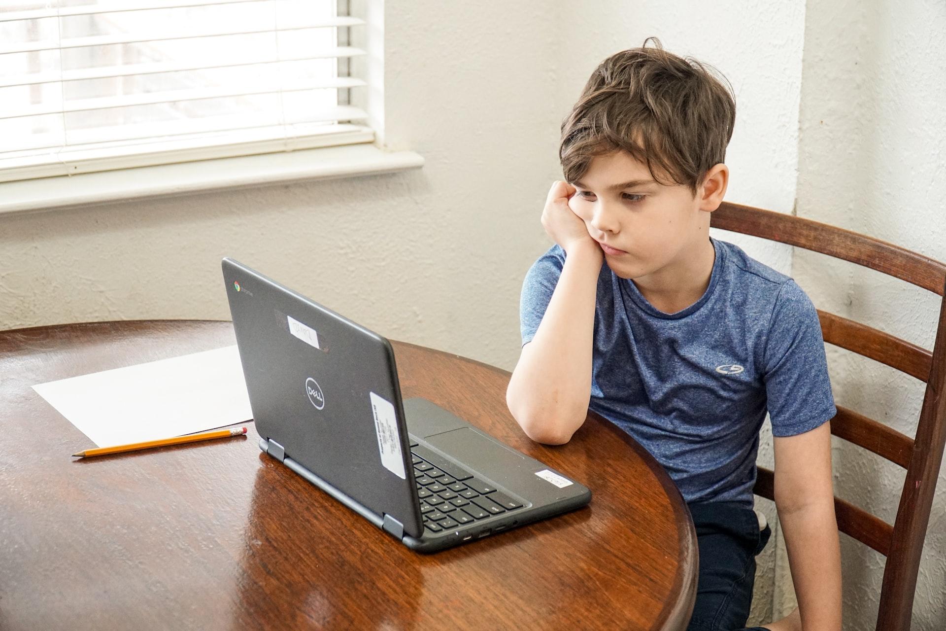 A boy in a blue shirt sits at a wooden table next to a window with his head propped up on his hand, looking bored as he gazes at an open laptop computer's screen. 