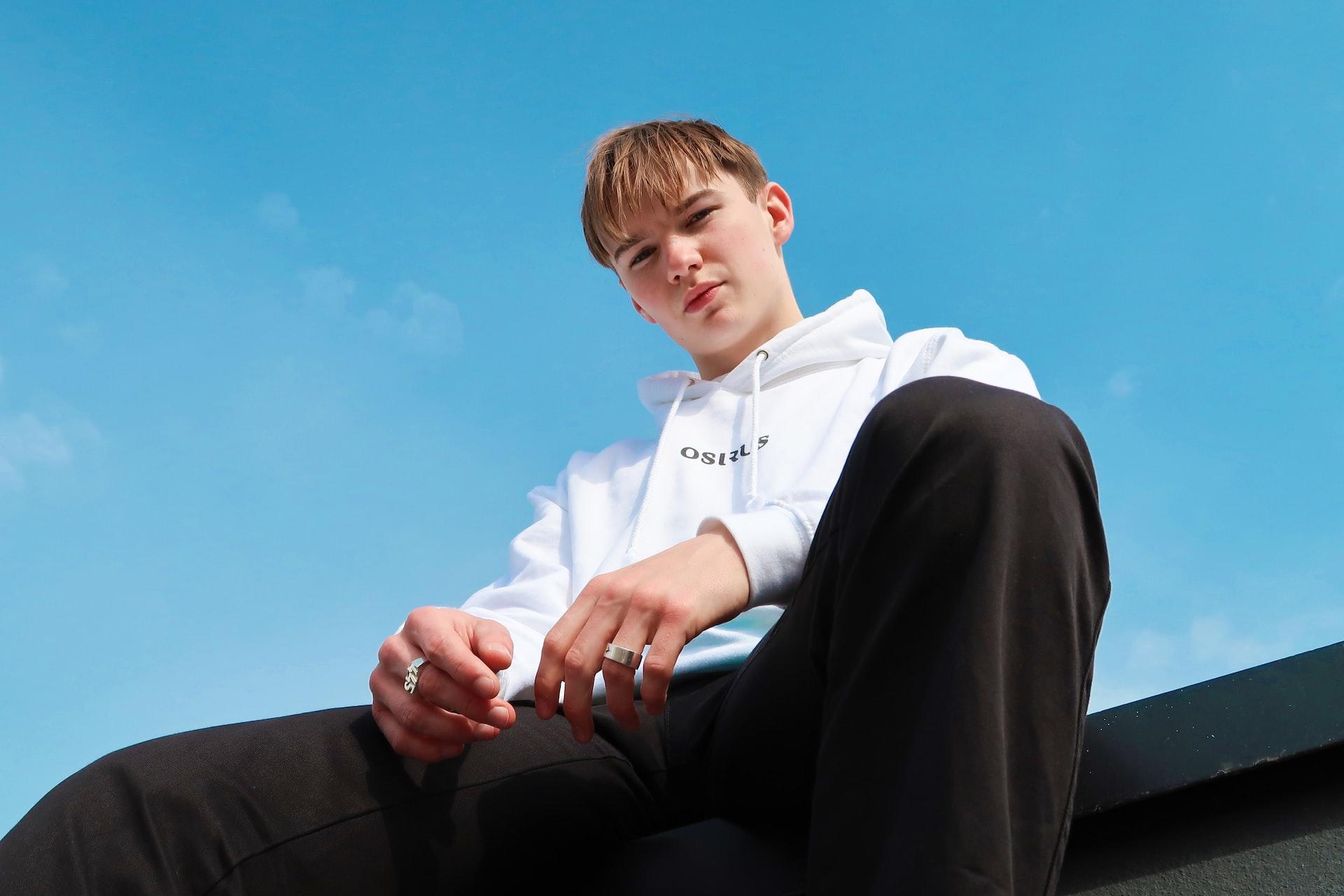 A teenaged boy wearing a white hoodies and black trousers sits up high, outlined against the blue sky, looking down at the camera.