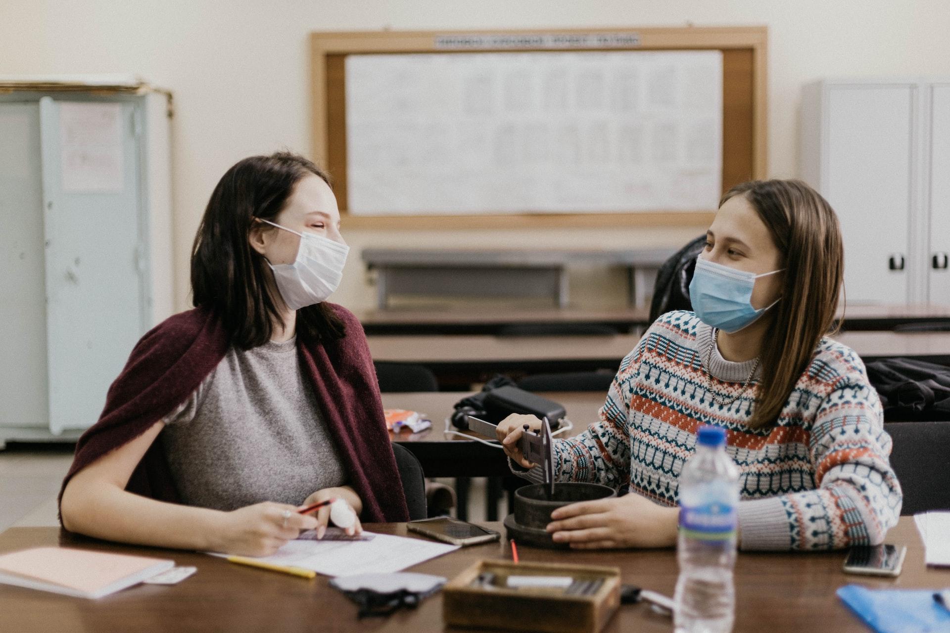 Two women wearing face masks sit in a classroom side by side, turned towards each other and smiling.