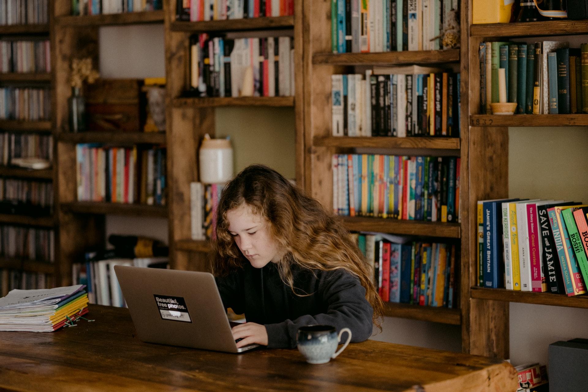 A girl with long hair sits in a home library, at a wooden table, looking at something displayed on her laptop computer's screen. 