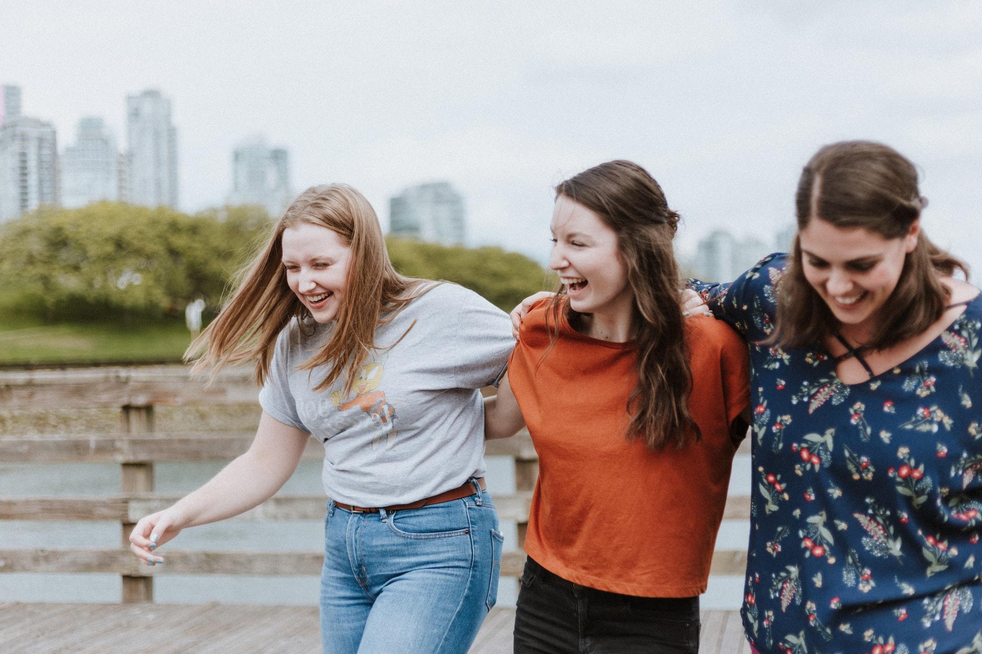 Three girls walking through the countryside arm in arm and laughing with a city skyline visible in the distant background. 