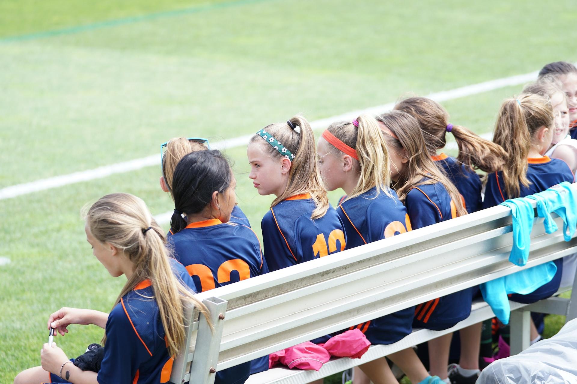 A group of pre-teen girls with their hair tied back, wearing sports uniforms, sits on a bench alongside a football pitch