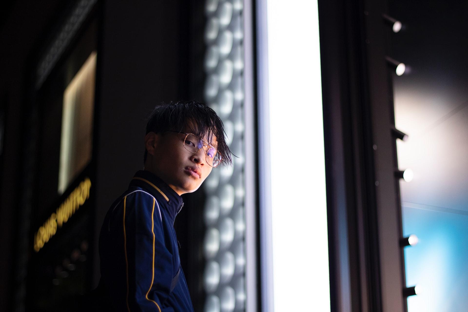 A young boy wearing a purple top with gold piping stands in a dark corridor, by a window that highlights his appearance. 