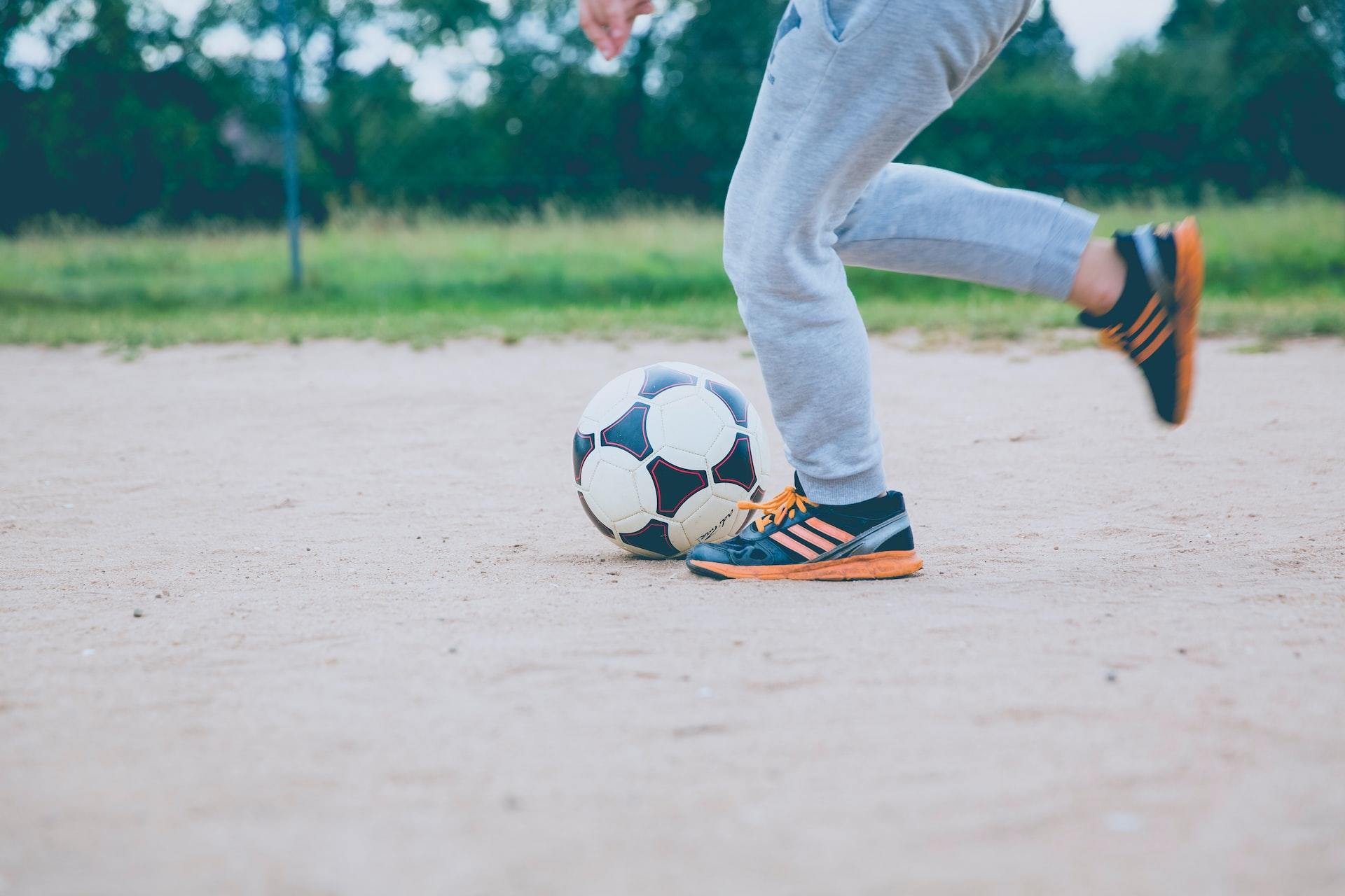 A person's legs shown from the mid-thigh down with the back leg raised, prepared to kick a black and white football. 