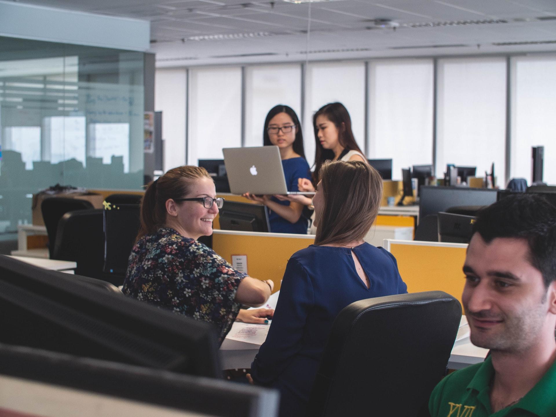 An office setting where two girls stand in the background with a laptop computer held up for them to read and a woman interviewing a student in the foreground