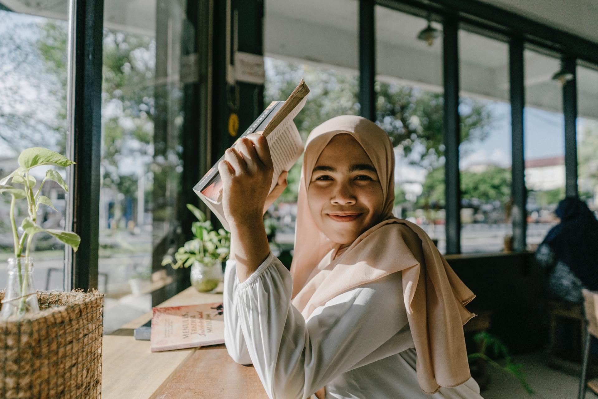 A girl in a pale-coloured hijab sits at a ledge table facing a bank of windows, holding a book up to shield her eyes from the sun and smiling