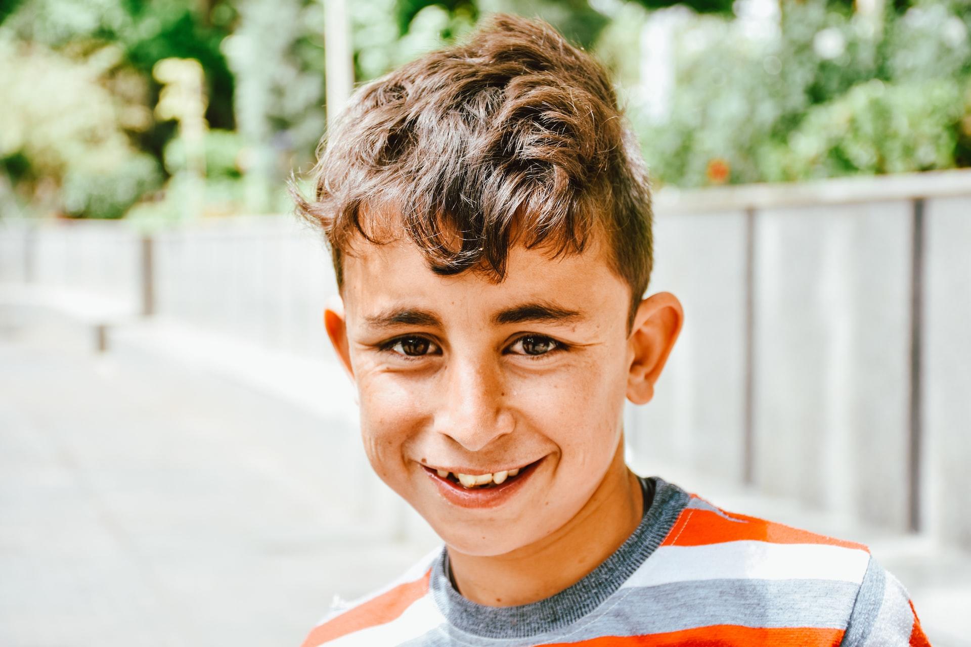 A boy in a striped orange blue and white shirt, with a tousled mop of hair, stands outside on a sunny day, smiling for the camera. 